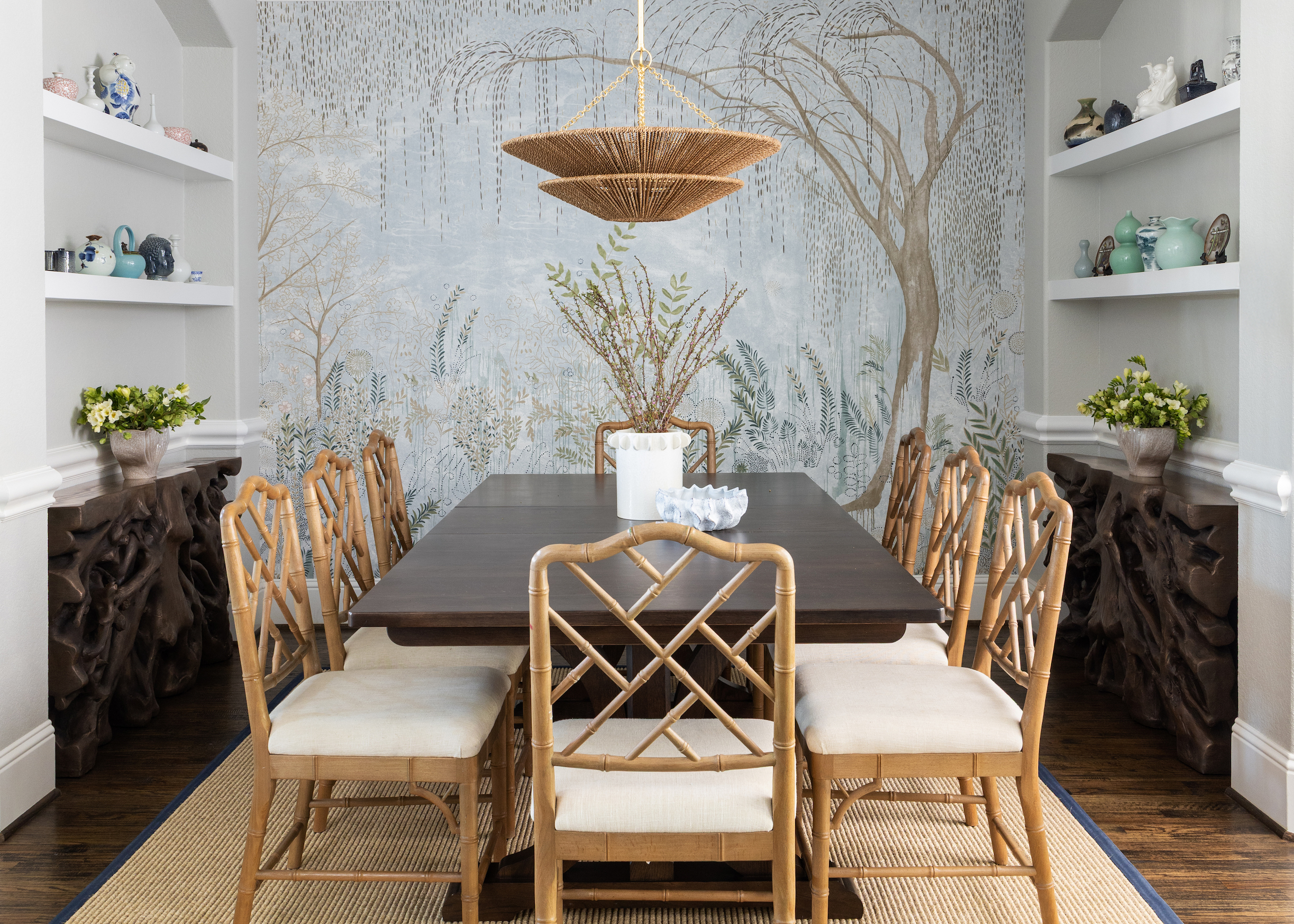 Dining room with a dark wood table, bamboo-style chairs, and a botanical mural backdrop. A woven pendant light hangs above, while built-in shelves showcase decorative vases and art pieces.