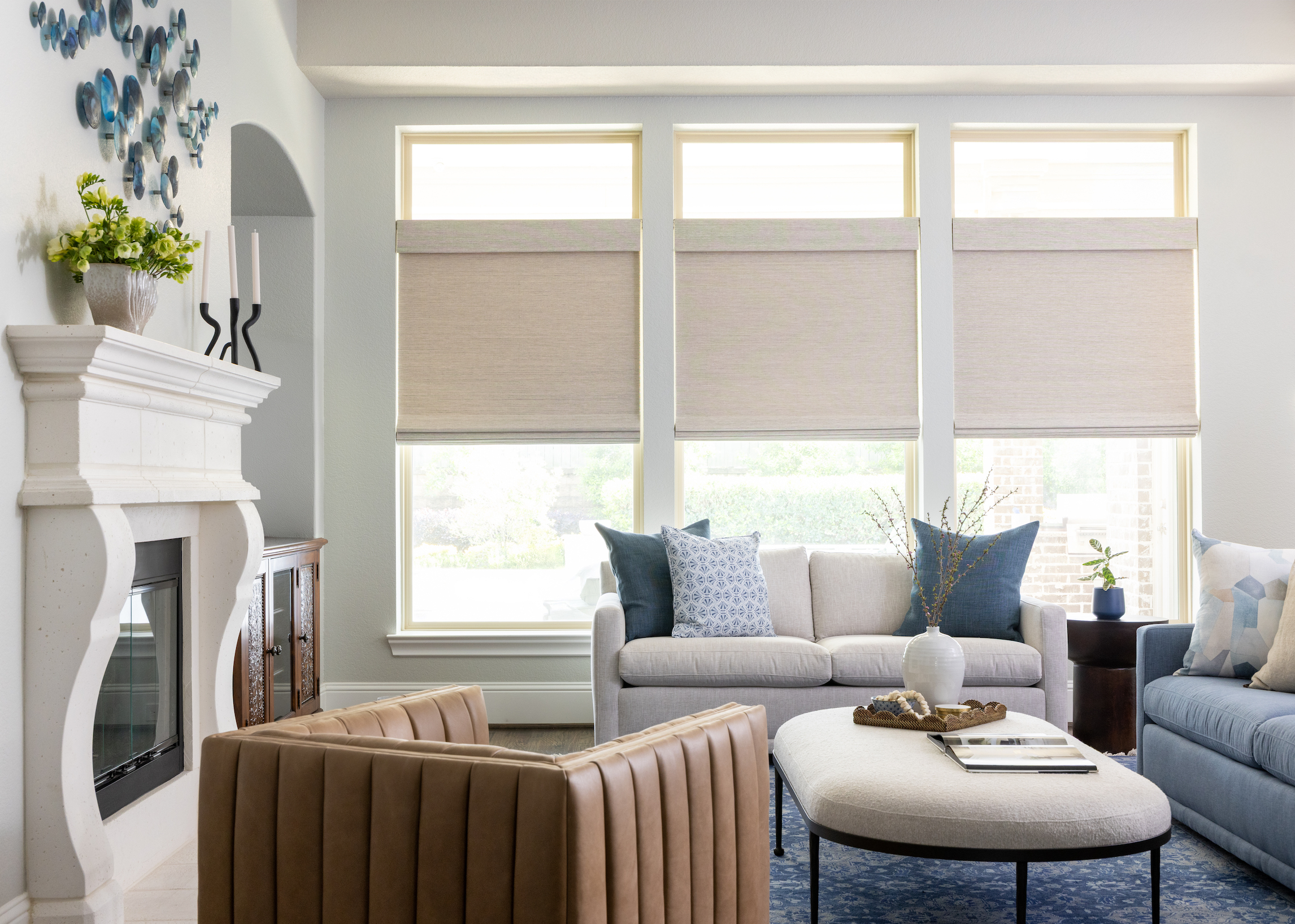 Bright living room with neutral sofas, blue accent pillows, and a tan leather armchair. Three large windows with beige Roman shades bring in light, and a soft ottoman coffee table sits in the center.