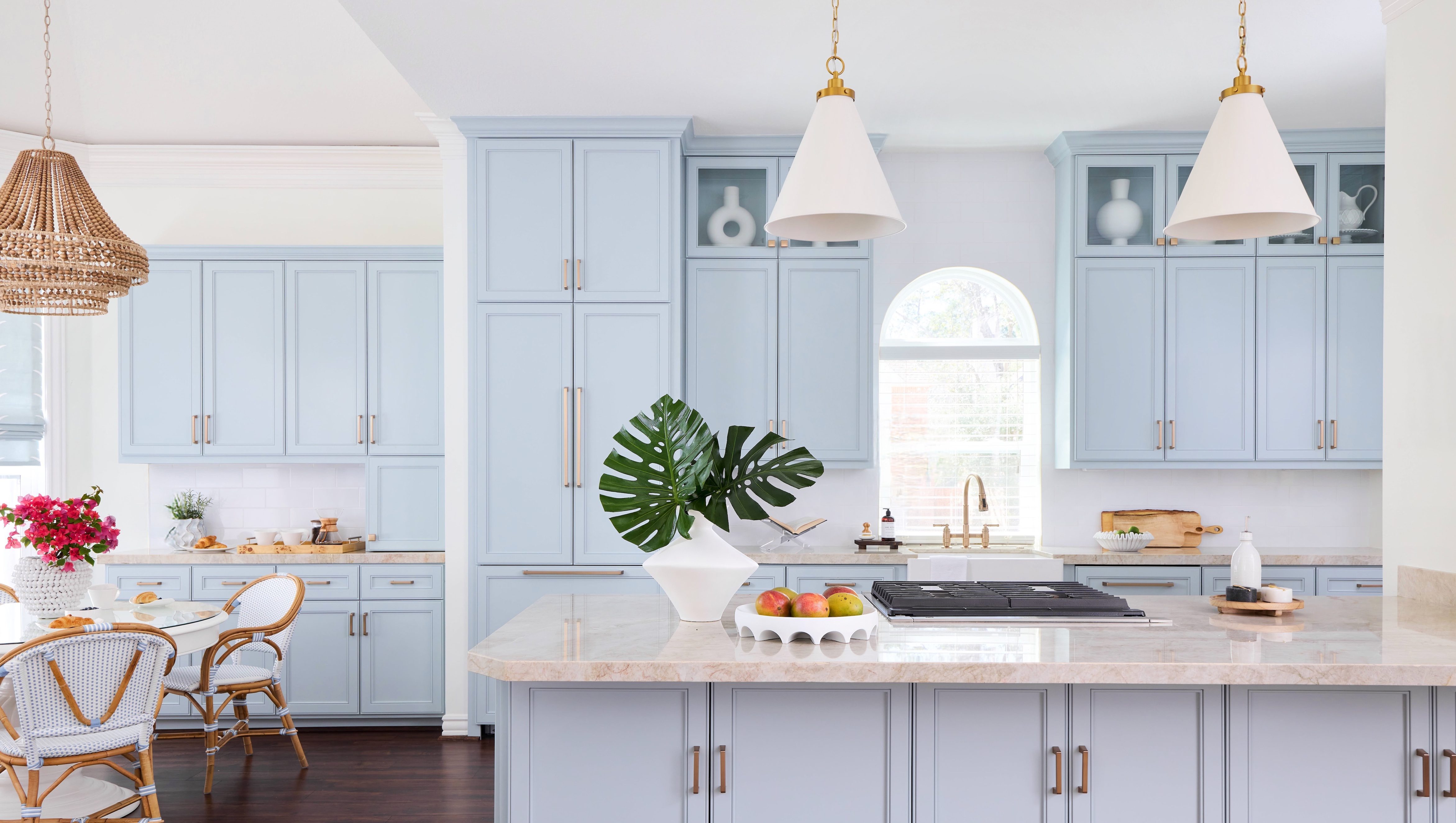 Spacious kitchen with pale blue cabinets, gold hardware, and a large marble island. Two white pendant lights hang above the island, which is decorated with a monstera leaf in a white vase and a tray of fresh fruit. A farmhouse sink and arched window bring in natural light, while a breakfast nook with wicker chairs is visible to the side.